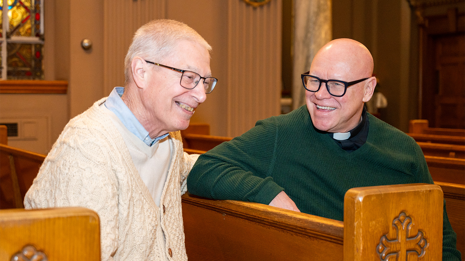 An older man and a priest sitting in pews in a hospital chapel, smiling and talking