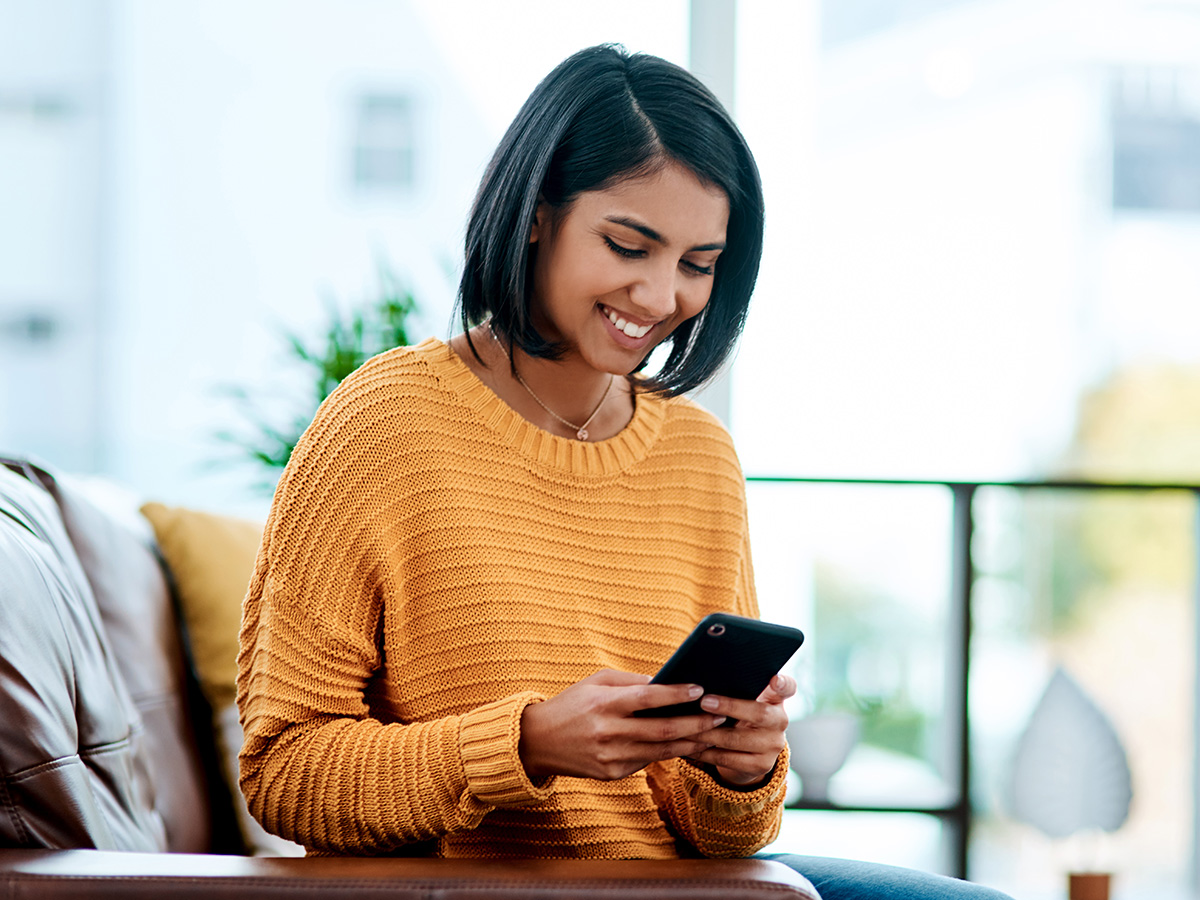 Woman with yellow sweater sitting on a couch and looking at her phone.