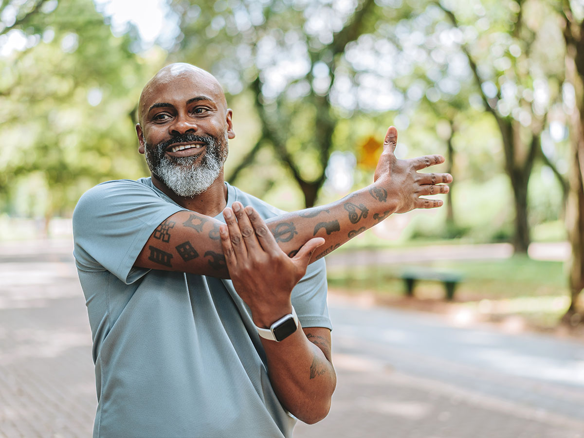 Smiling middle-aged man, stretching his arm across his chest on an outdoor paved path with trees