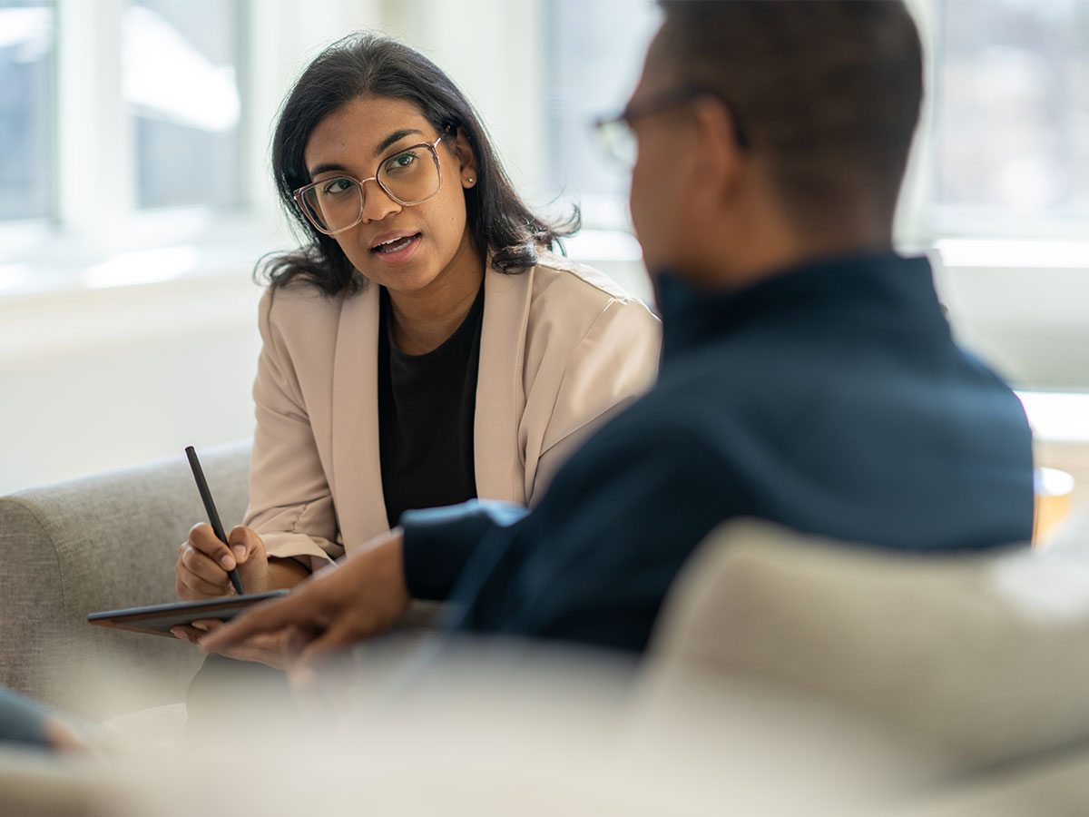 Woman in blazer sitting on couch taking notes and speaking to man in foreground