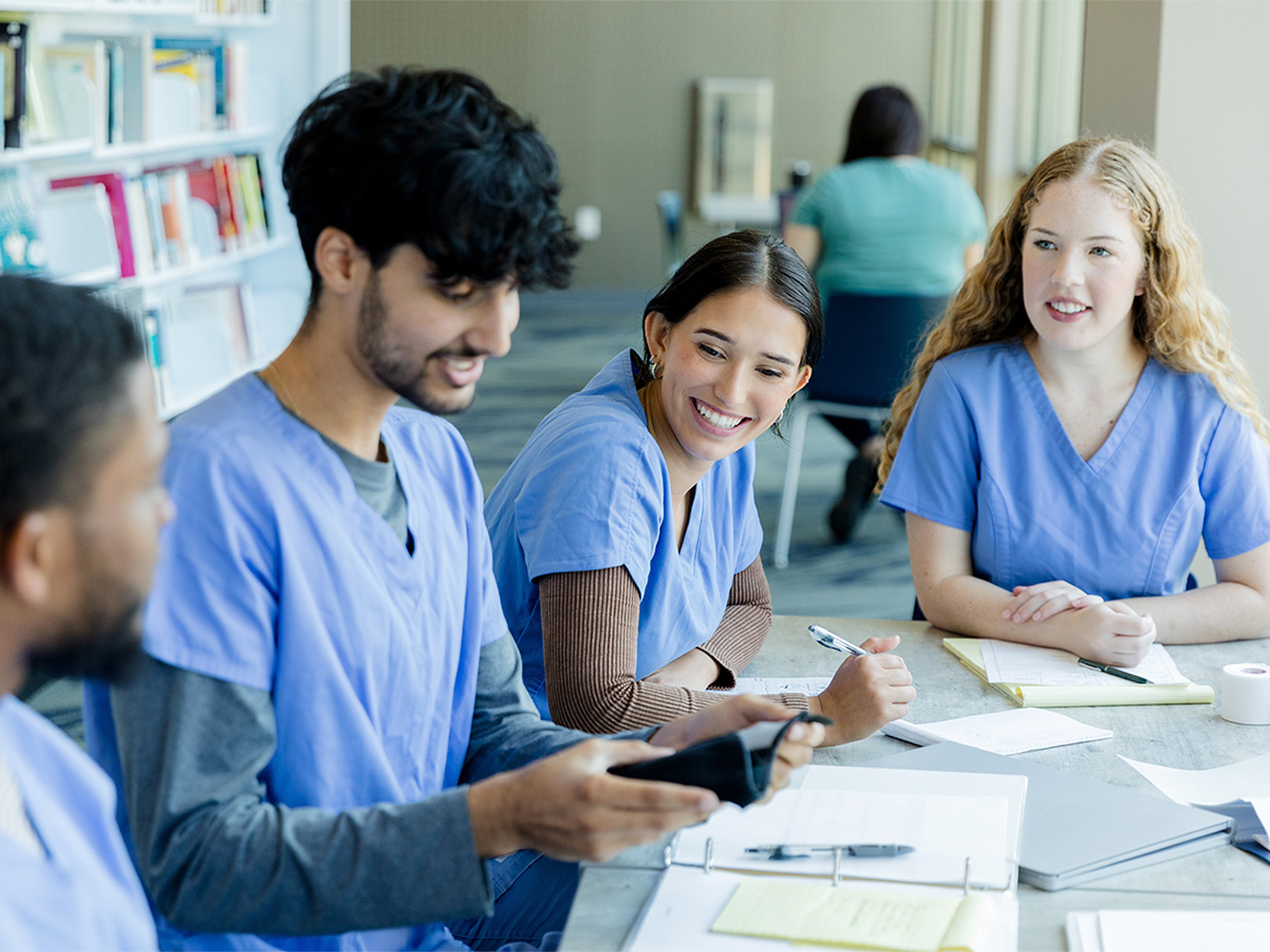 Two male and two female medical students, wearing blue scrubs, sitting at a table and working on an assignment
