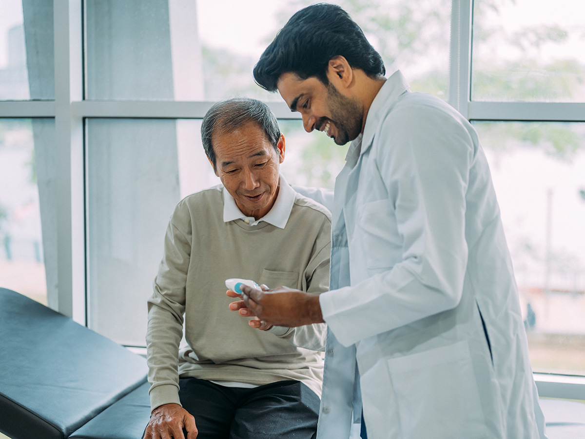 Young male physician in white lab coat shows plastic medical item to older male patient who is seated on table