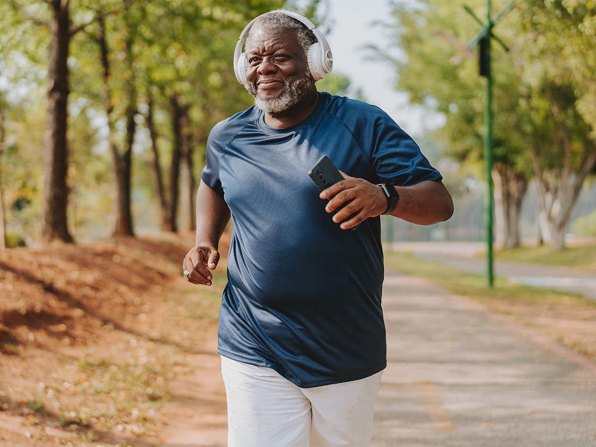 Older man, slightly overweight, wearing headphones, jogging through park on a paved trail lined with trees