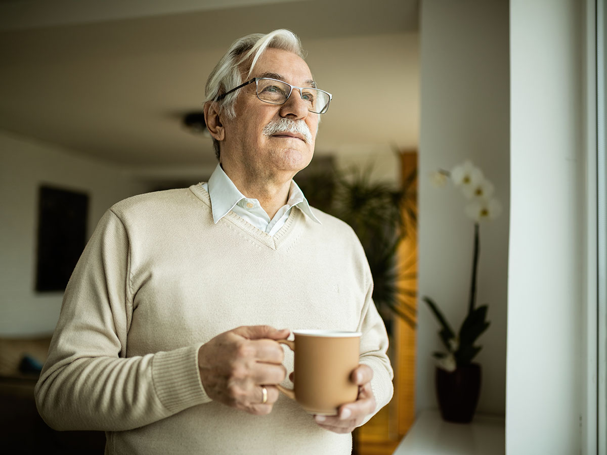 Adult male wearing tan sweater holding a coffee mug while looking through a window.