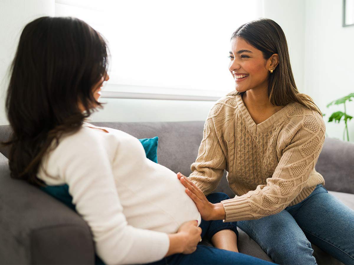 Pregnant woman on couch talking with her female friend, who has her hands on her belly