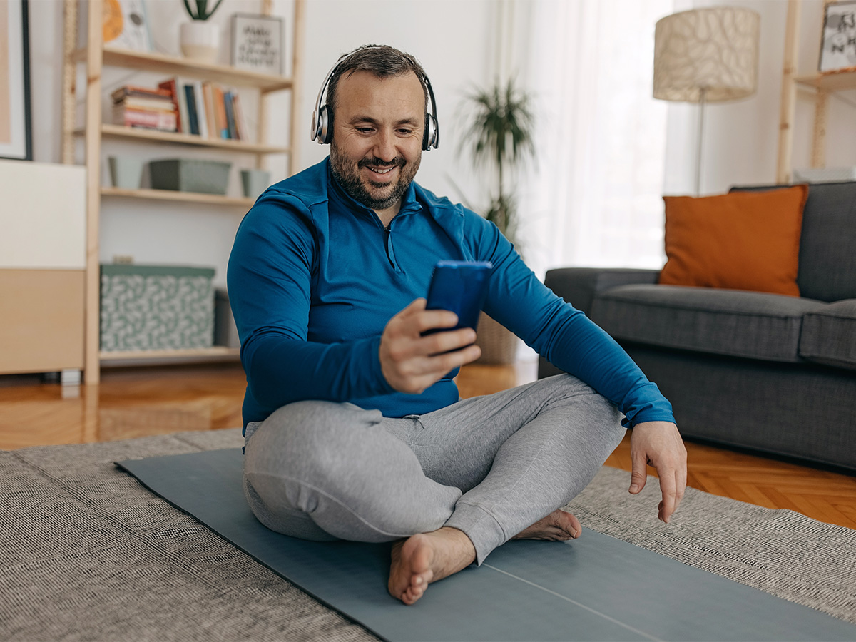 Overweight, middle-aged male sitting cross-legged on yoga mat, smiling and looking at his phone while wearing headphones