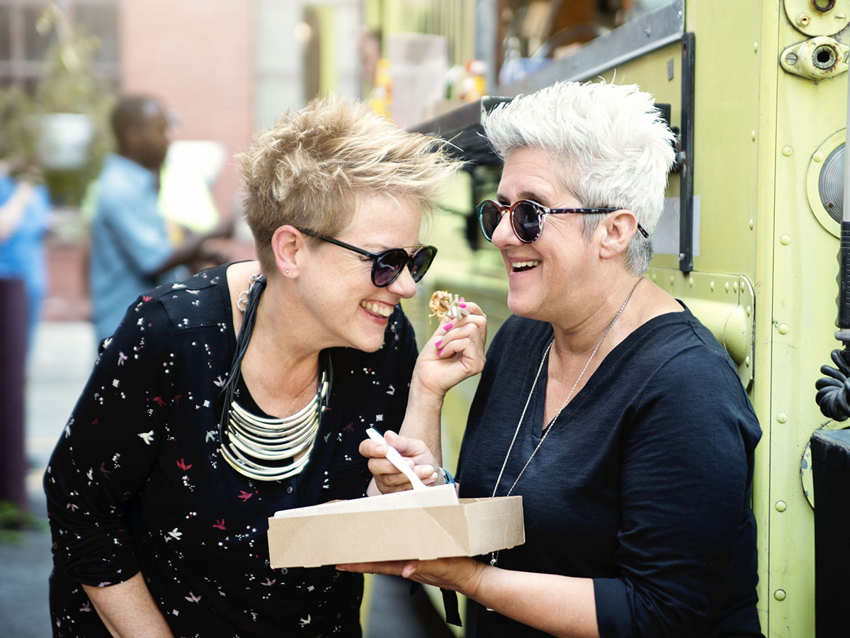Two older women laughing while eating lunch from a food truck