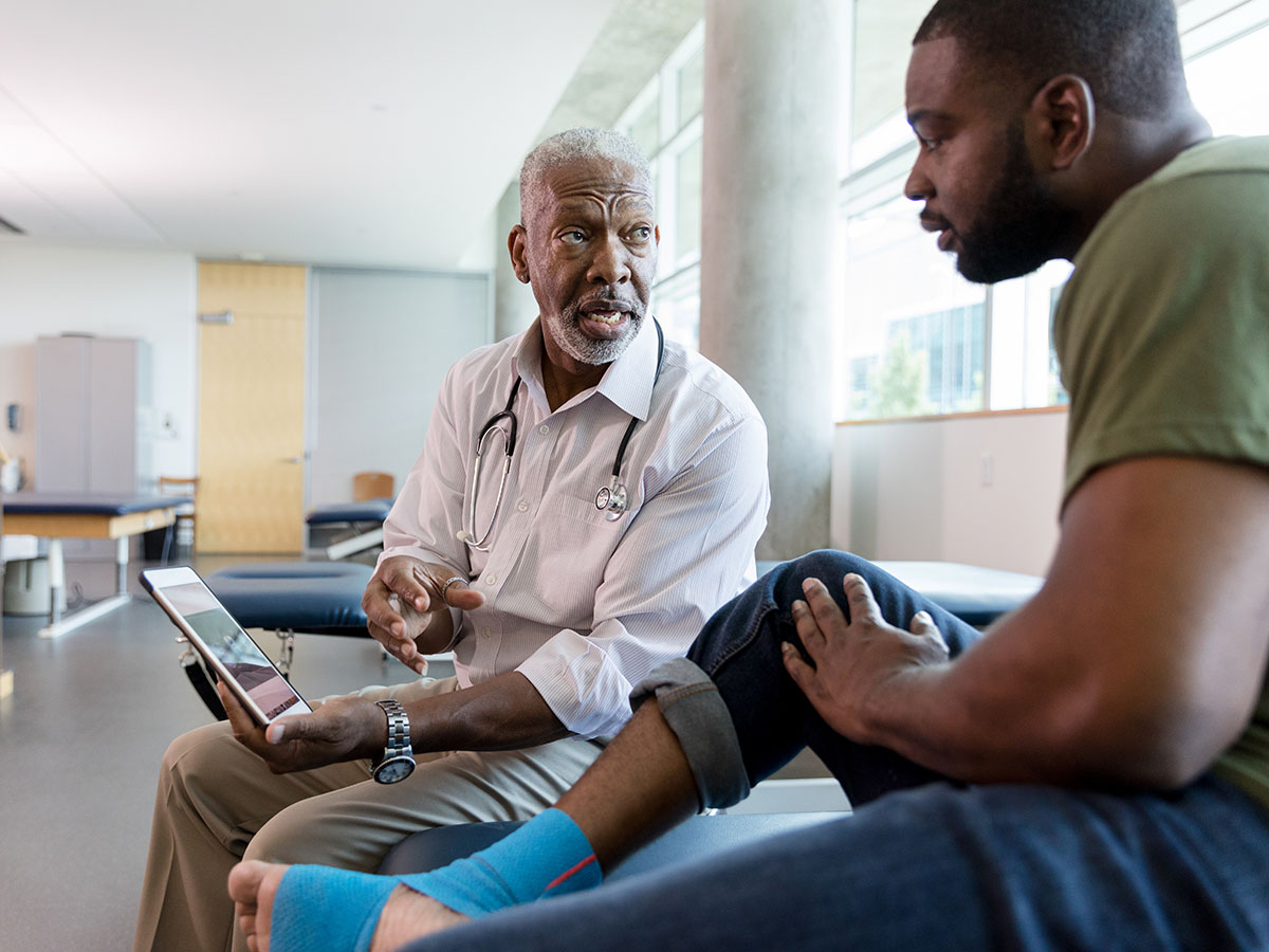 older male doctor showing ipad to younger male patient whose food and ankle are wrapped.