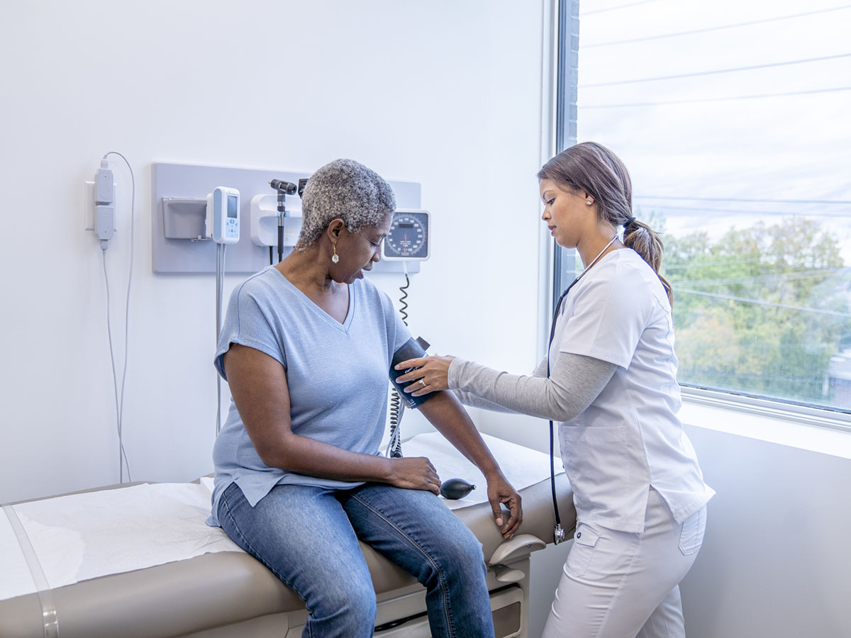 Female nurse in white scrubs checks blood pressure of an older adult female patient.