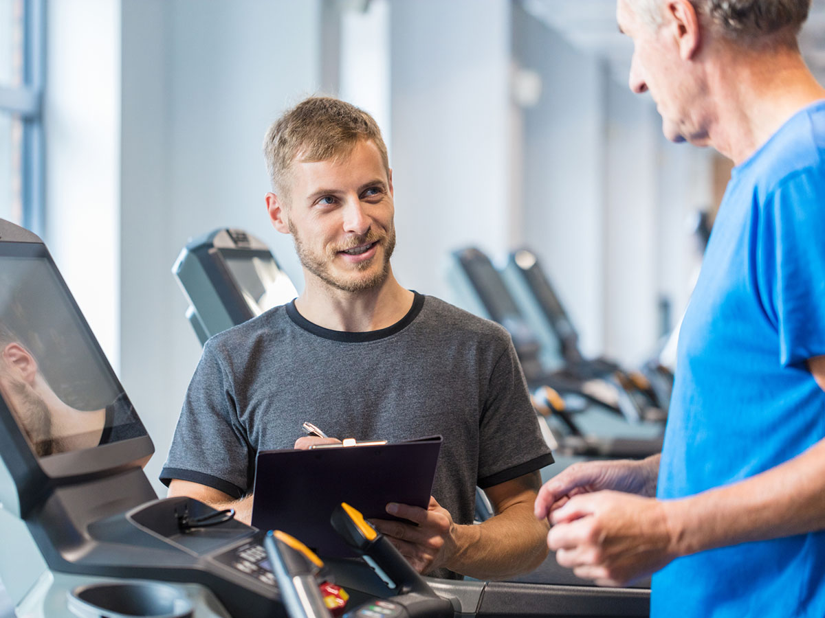 An older adult male in blue t-shirt is coached by a gym trainer on the treadmill.