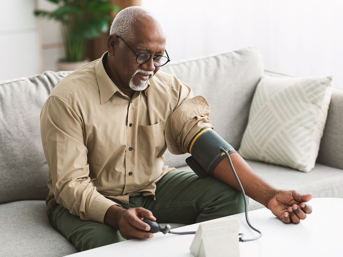 An older male adult in tan button down shirt checks his blood pressure.