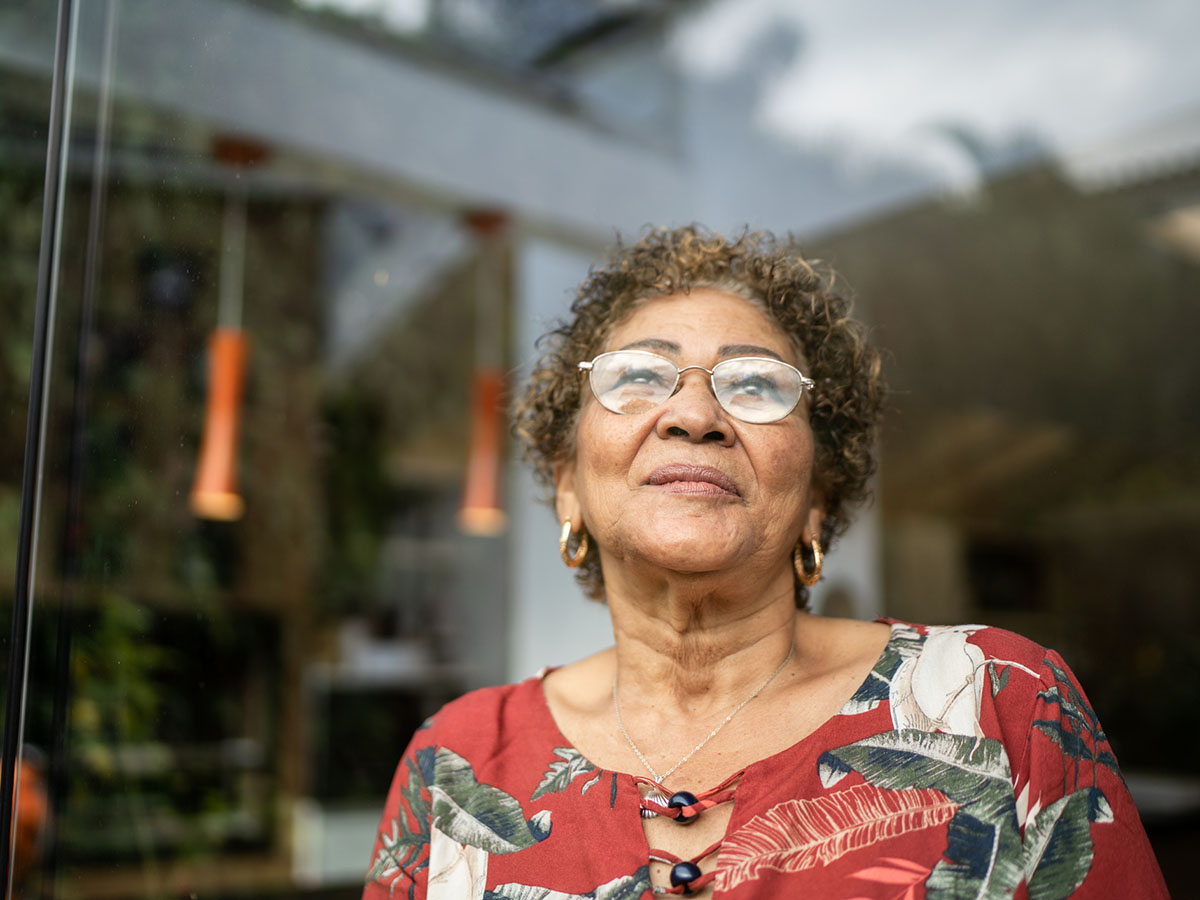 Older woman in floral shirt and glasses stands outside her house.