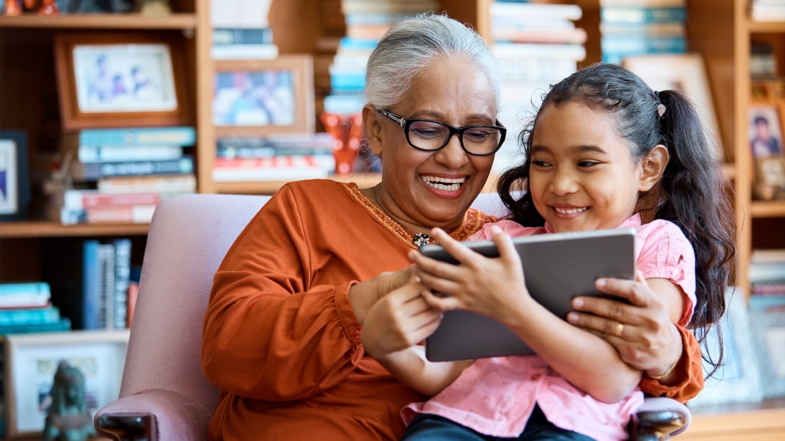 A grandmother and granddaughter sit in a chair and watch a tablet