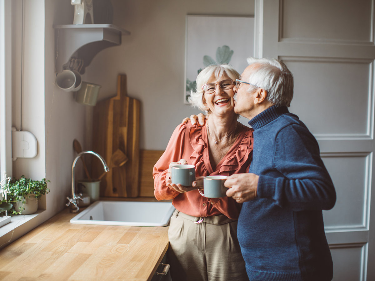 Older couple hold coffee mugs and embrace in the kitchen.
