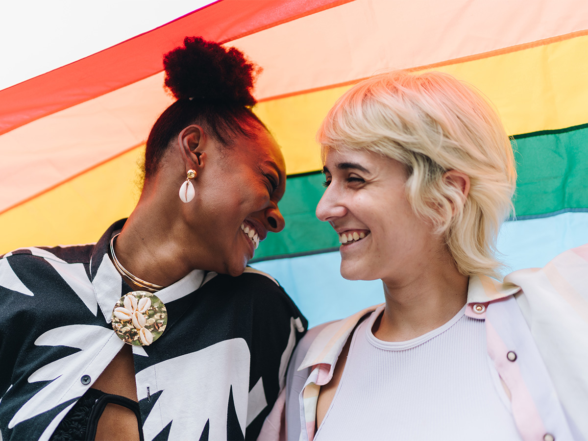 Young black woman smiling at young white woman in front of a painted backdrop resembling a rainbow