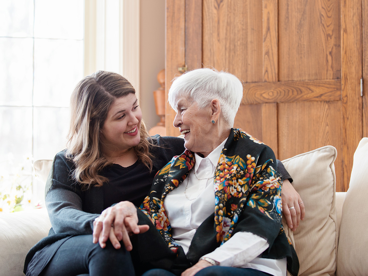 Adult woman and elderly woman embrace on a couch.