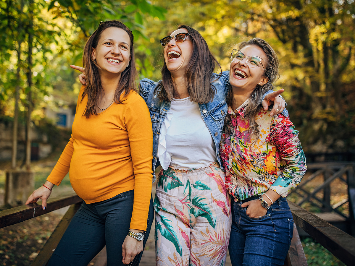 Three women of varying ages laughing and walking in park, one woman is pregnant