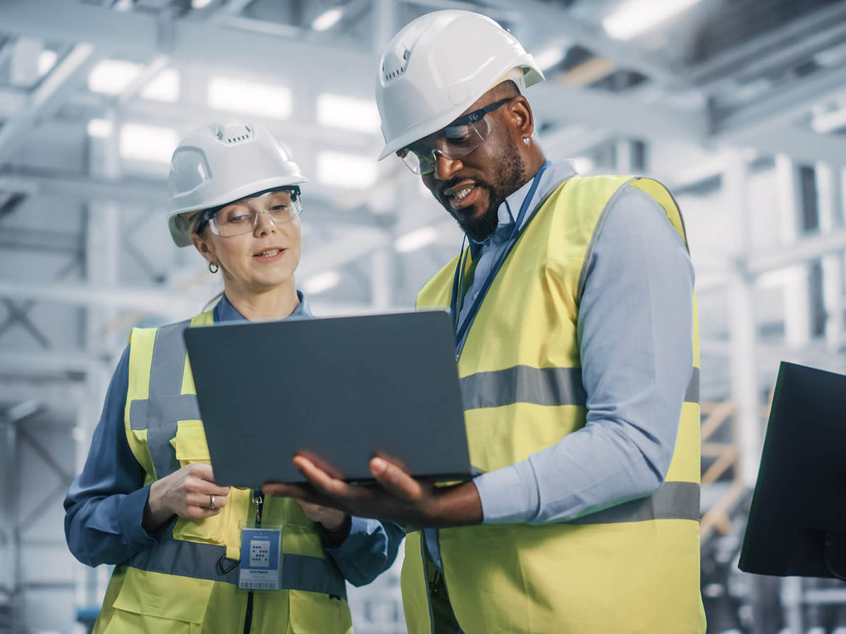 Man and woman in hard hats standing on a factory floor and looking at laptop screen
