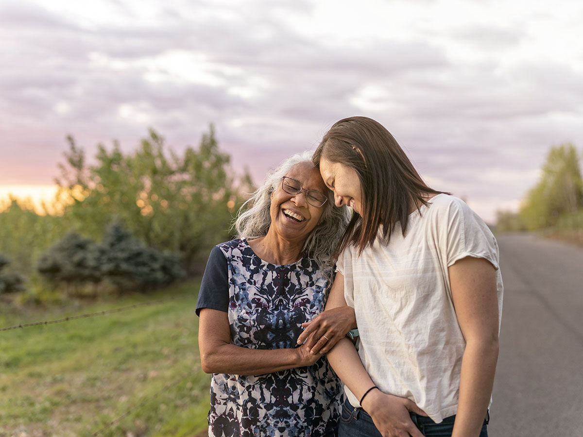 Older women adult and her younger daughter out for a walk before the sun sets, smiling and holding arms 