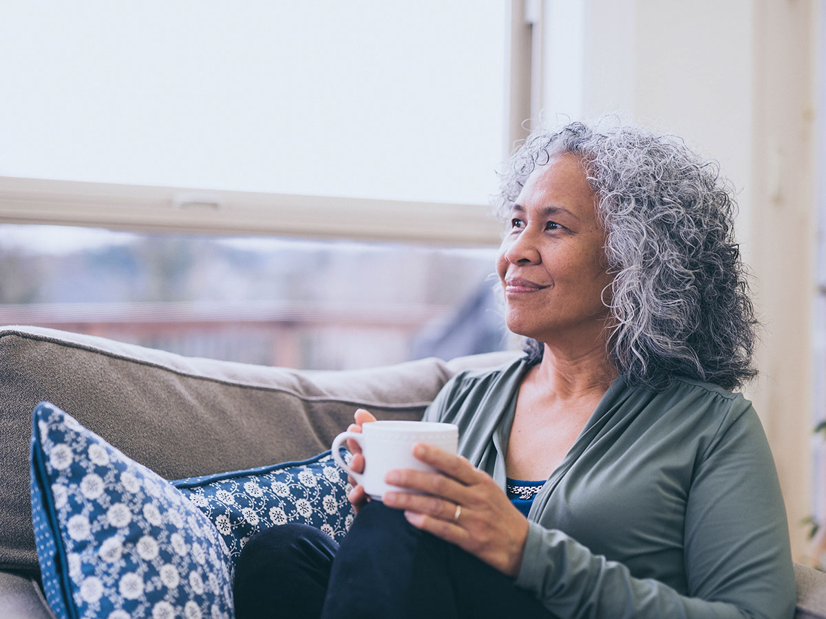 A senior woman on the couch holding a coffee mug while looking out of a window.
